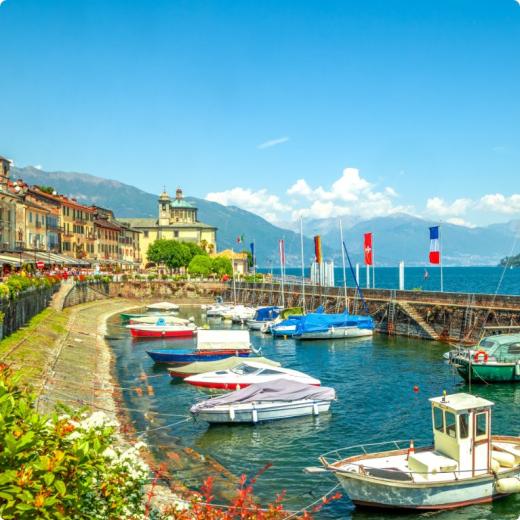 Porticciolo con barche colorate, edifici storici e montagne sullo sfondo in una giornata soleggiata.