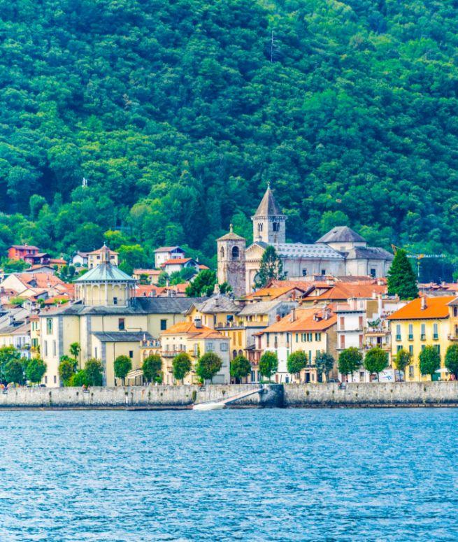 Colorati edifici sul lago, con colline verdi e una chiesa sullo sfondo.